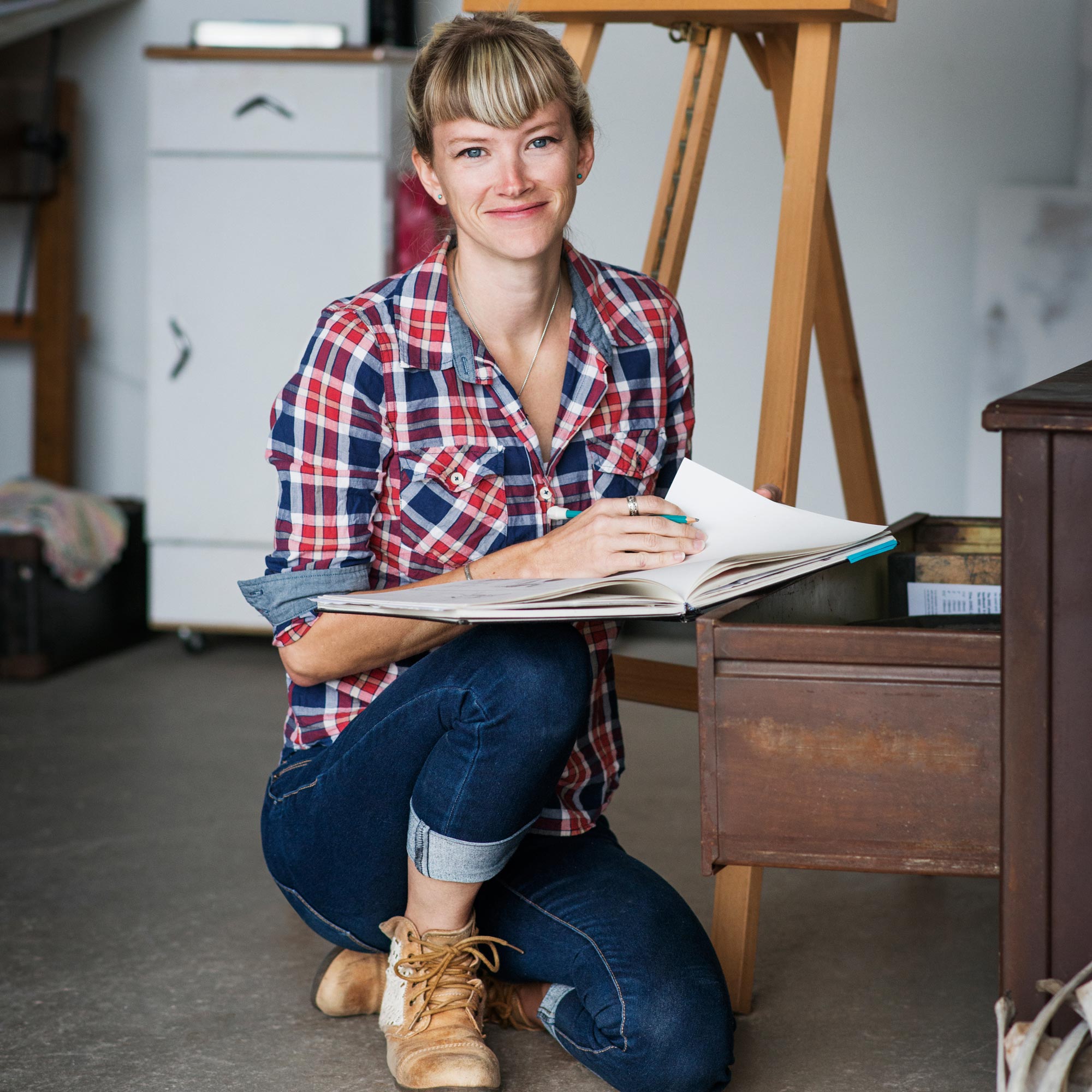 Leende kvinna med skjorta och jeans sitter på huk i ett rumm och bläddrar i en bok